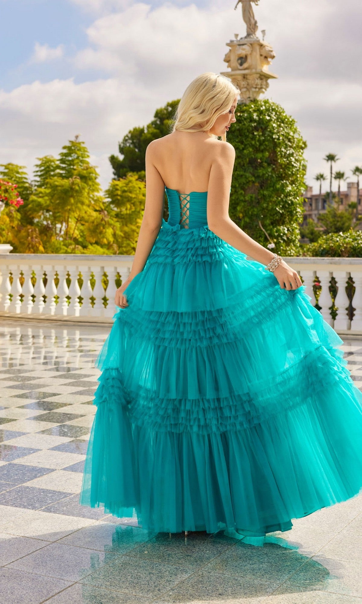 A woman in the Juliet JT2542S long prom dress with ruffled, layered turquoise fabric stands on a tiled terrace, holding her gown and facing away, with greenery and a statue in the background.